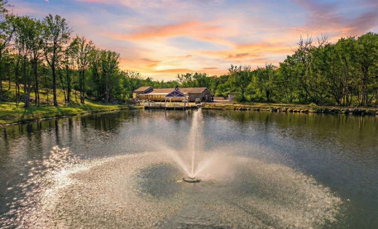 Lake fountain spraying water at sunset with a house and trees on the shore