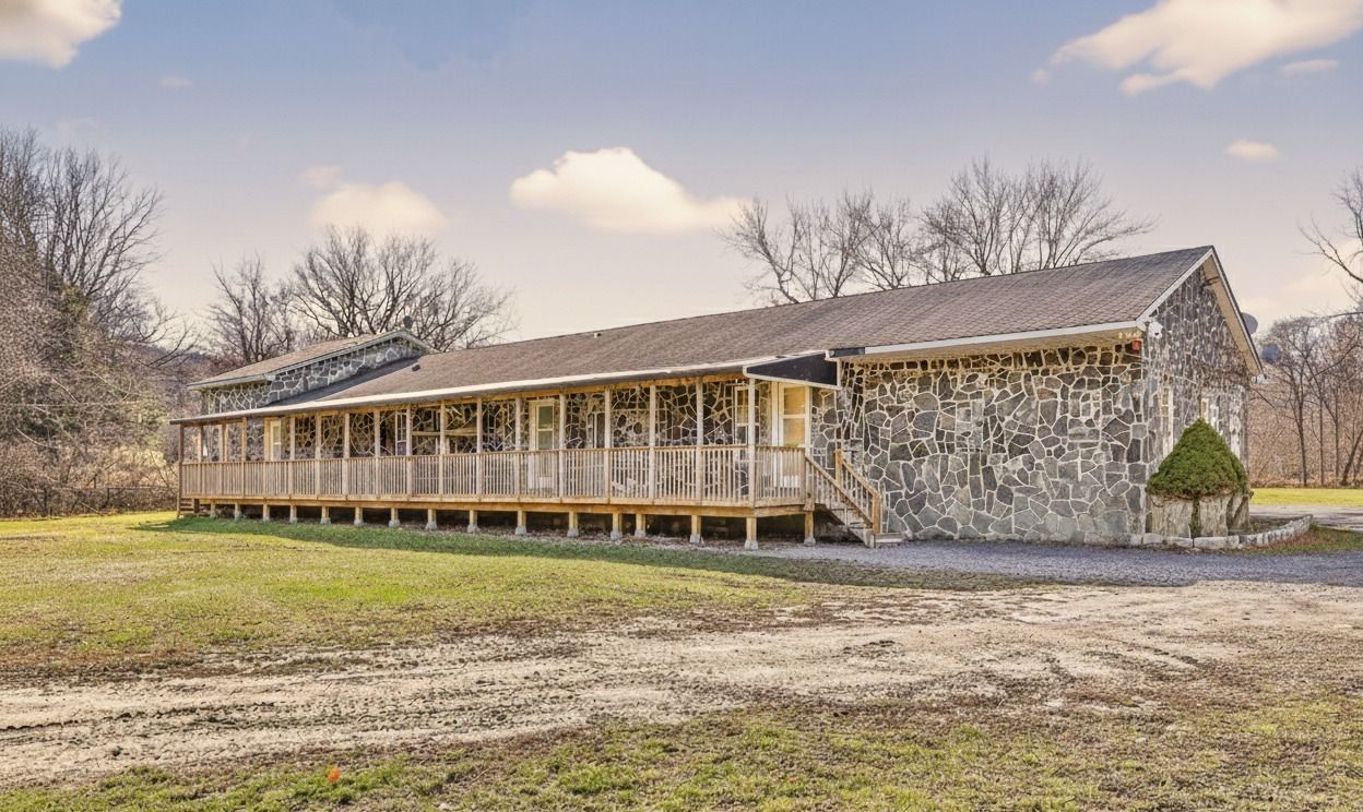 Long, weathered wooden building with a porch in a grassy field under a partly cloudy sky
