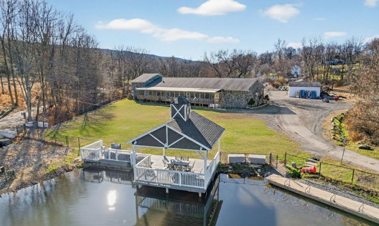 Aerial view of a dock, boathouse, and calm waterfront beside a gravel lot and bare trees