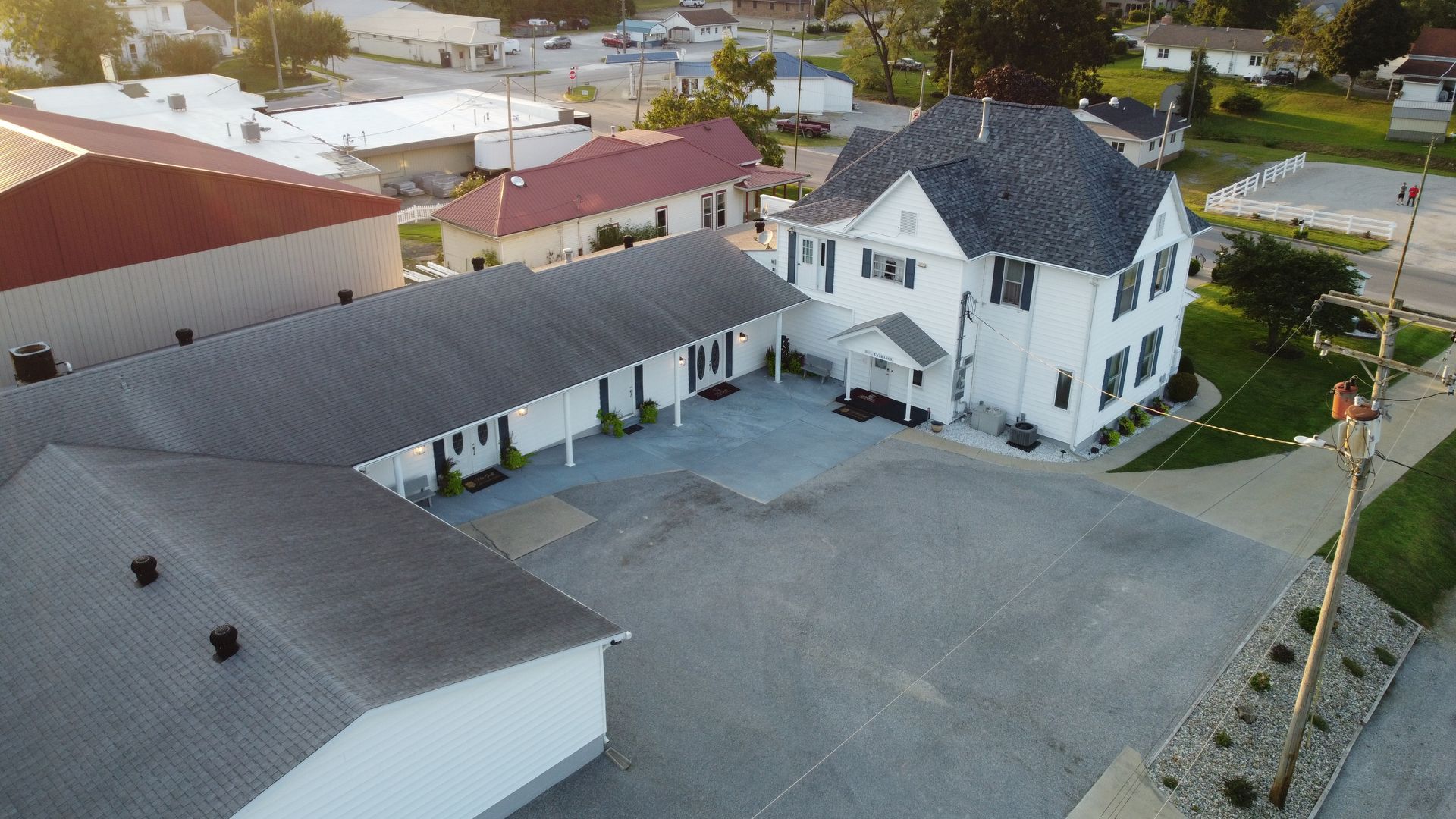 Aerial view of a white two-story building with a large gray roof beside a paved lot and nearby houses.