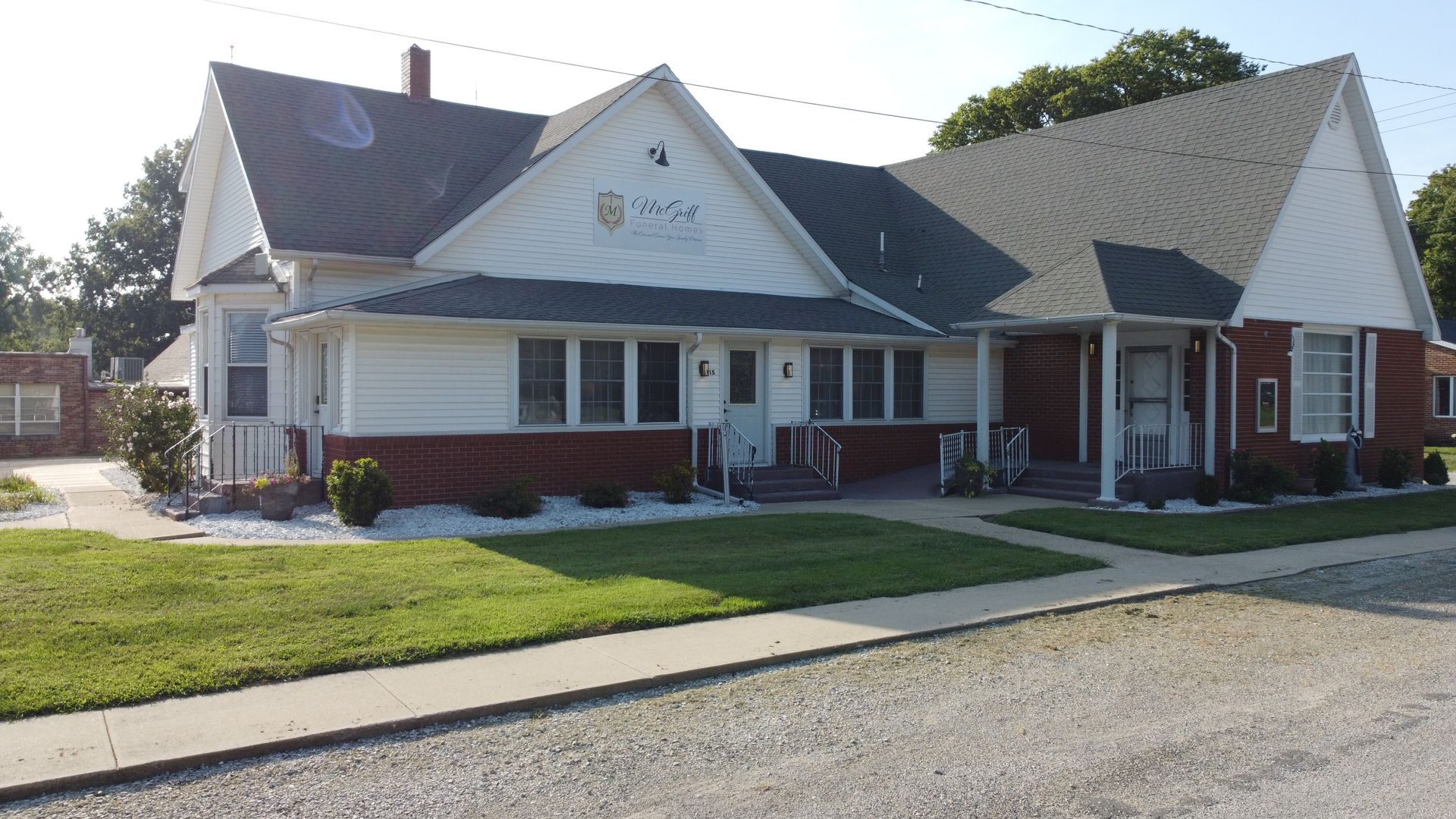 Brick-and-white community building with a peaked roof, lawn, and driveway on a sunny day