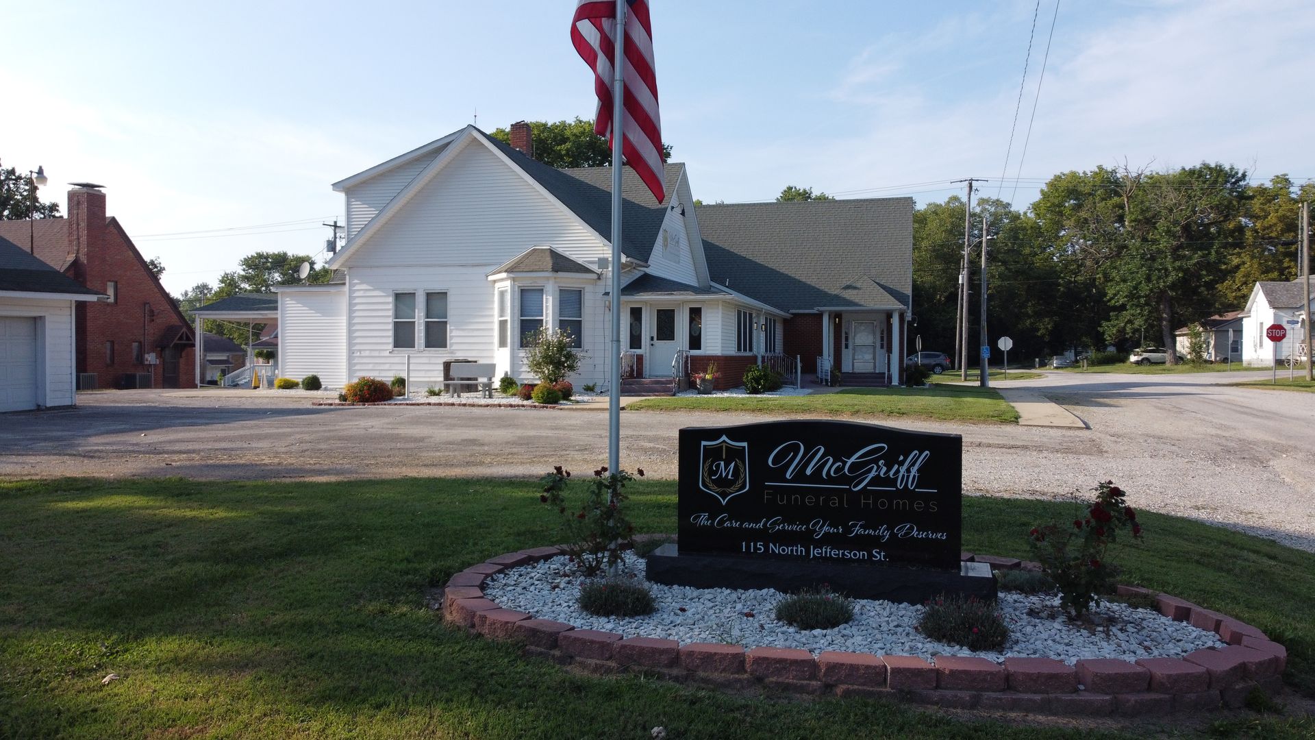 White civic building with an American flag and a landscaped sign in front.