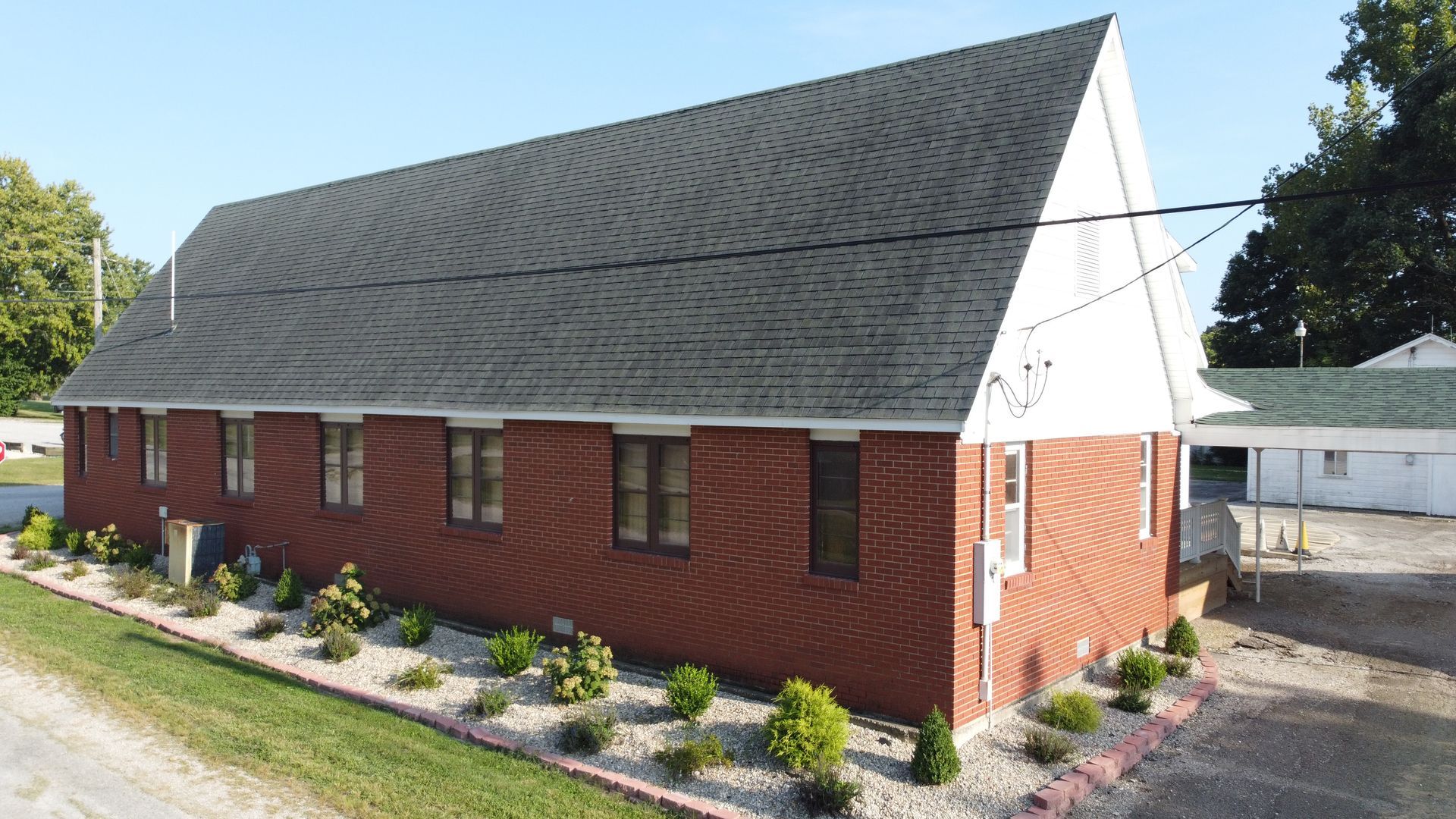 Red brick church with a steep gray roof, white trim, and landscaped flower beds beside a driveway