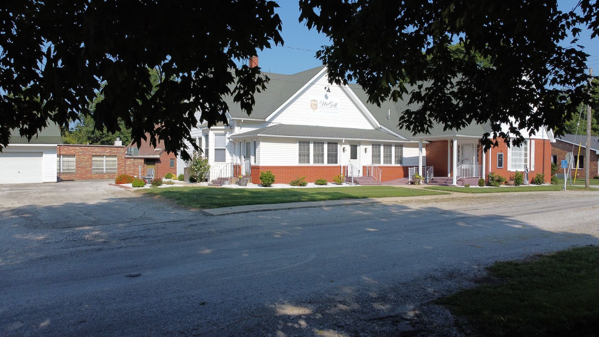 Small white and red brick building with a gabled roof, viewed across a paved parking lot under trees.