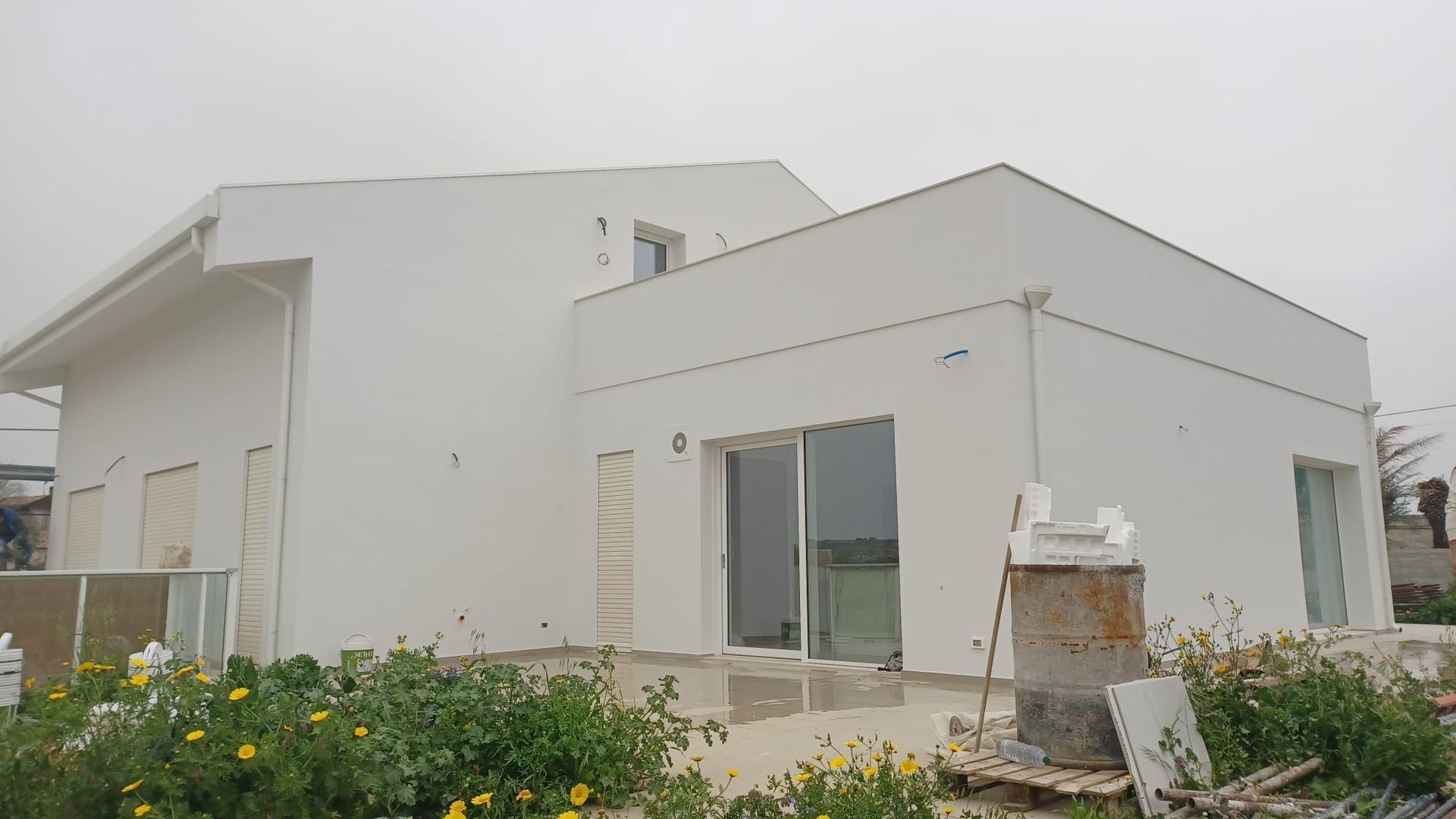 Casa moderna bianca con tetto spiovente e porta scorrevole in vetro, circondata da piante sotto un cielo nuvoloso.