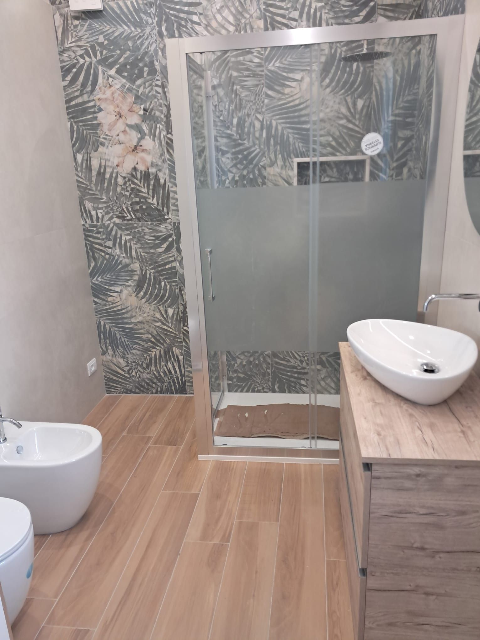 Bagno moderno con doccia in vetro, parete decorata, pavimento in legno e lavabo bianco su mobiletto in legno.