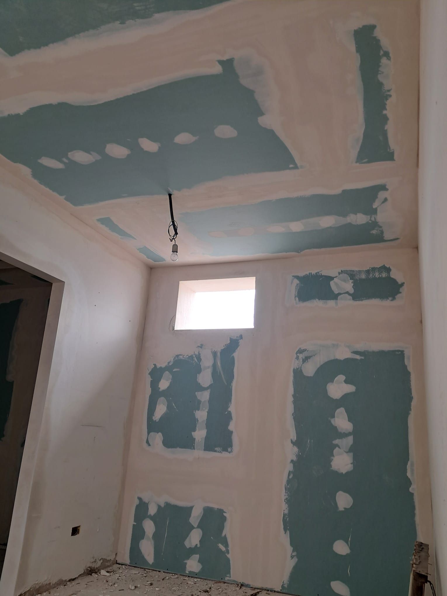 Stanza parzialmente rifinita con toppe di cartongesso grigio-blu, una piccola finestra e una lampada a sospensione.