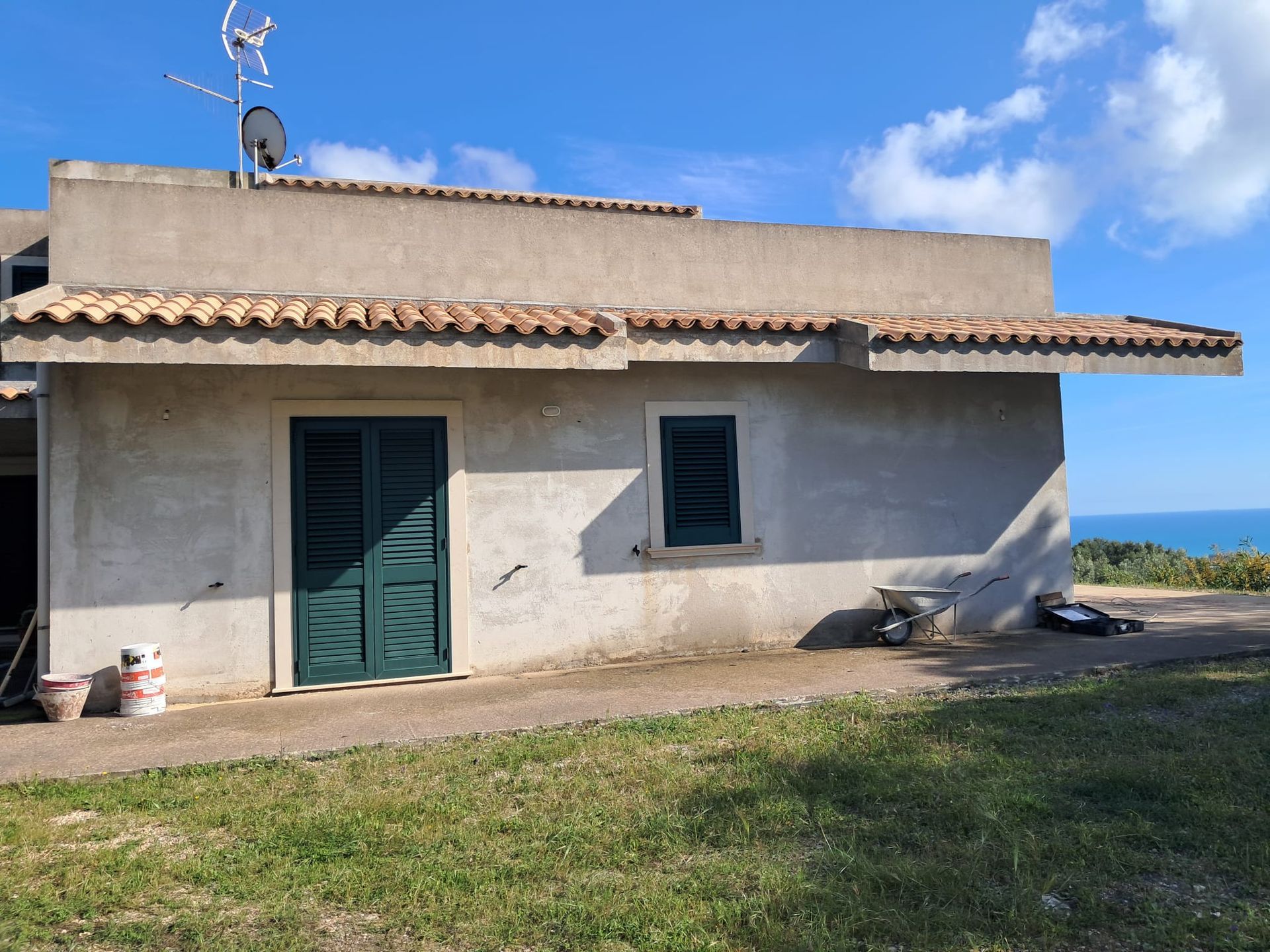 Piccola casa in stucco con persiane verdi e tetto in terracotta, situata accanto a un prato e all'orizzonte blu del mare.
