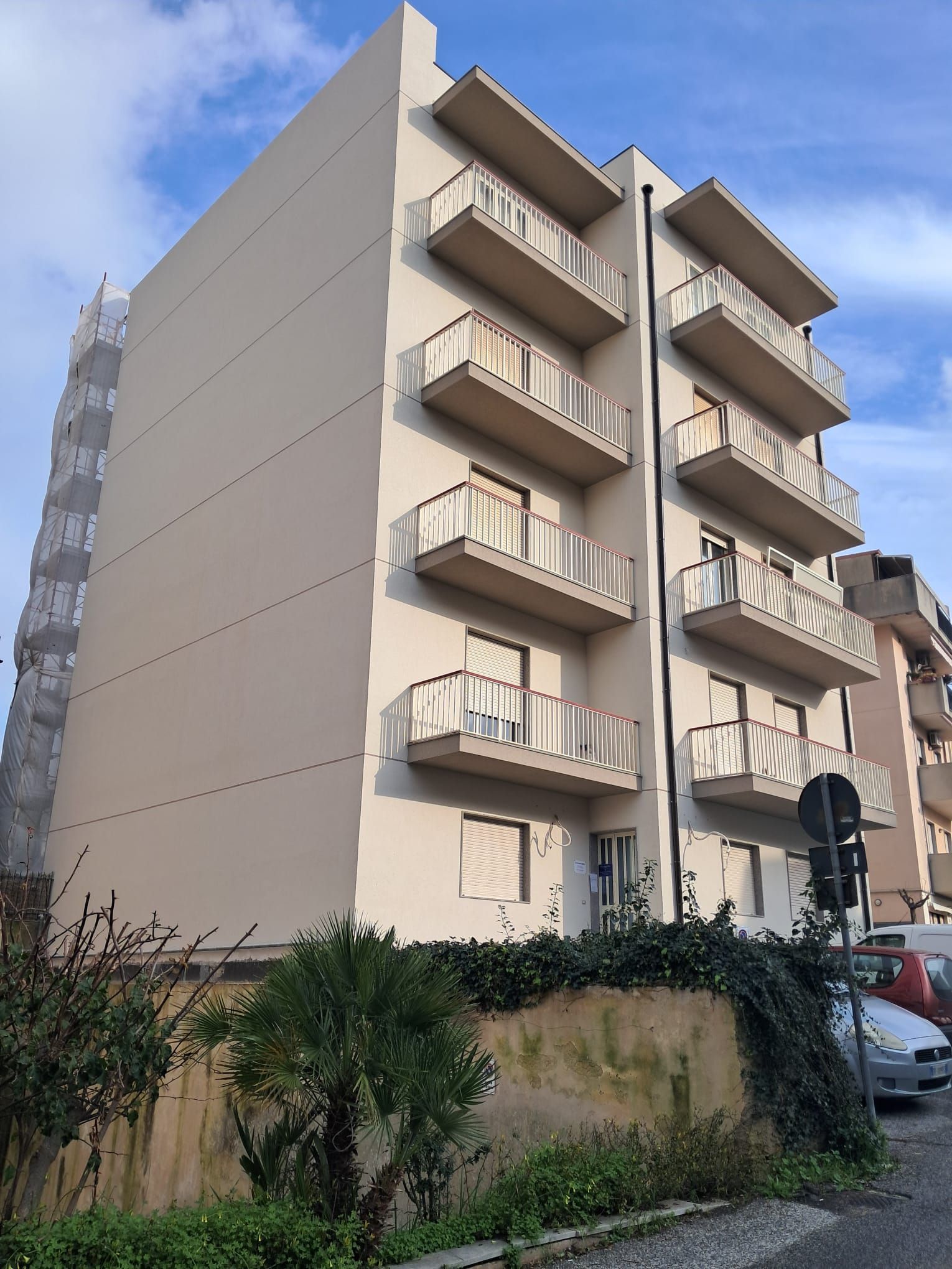 Edificio residenziale beige di cinque piani con balconi, visto dal basso tra aree verdi e auto parcheggiate.