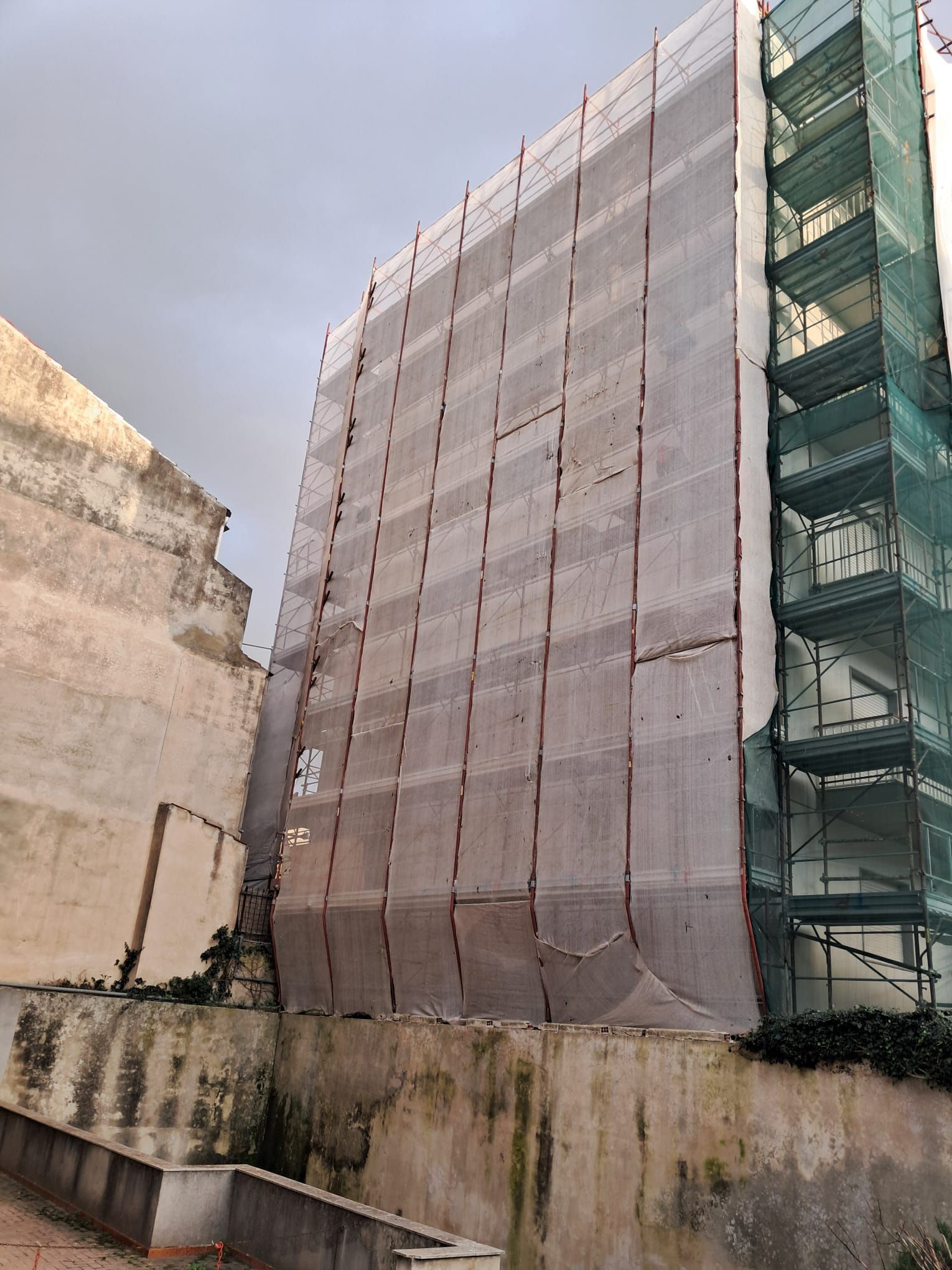 Un alto edificio in costruzione, avvolto in una rete protettiva, si erge accanto a un vecchio muro di cemento e a un'impalcatura per scale.