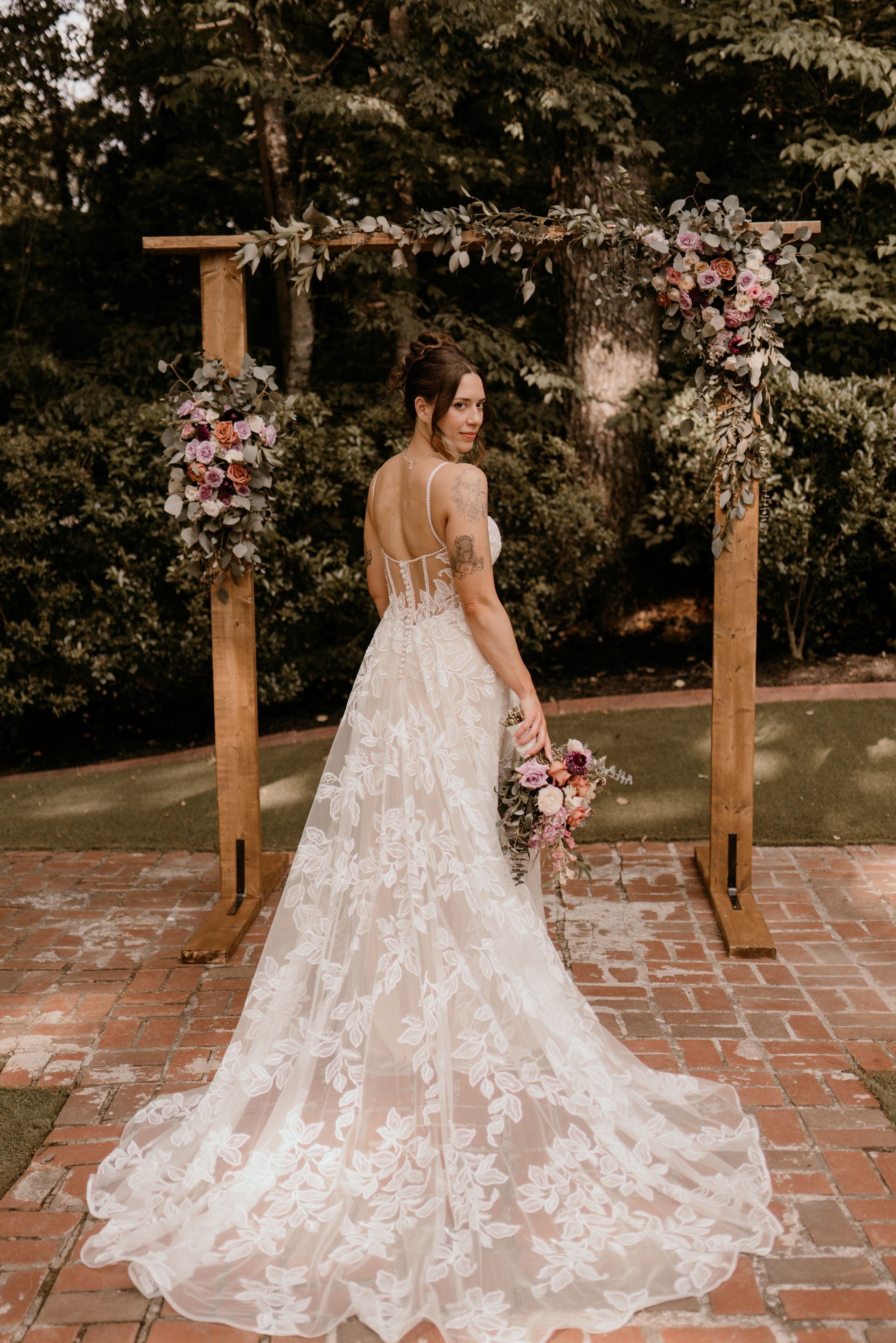 A bride in a wedding dress is standing in front of a wooden arch holding a bouquet of flowers.