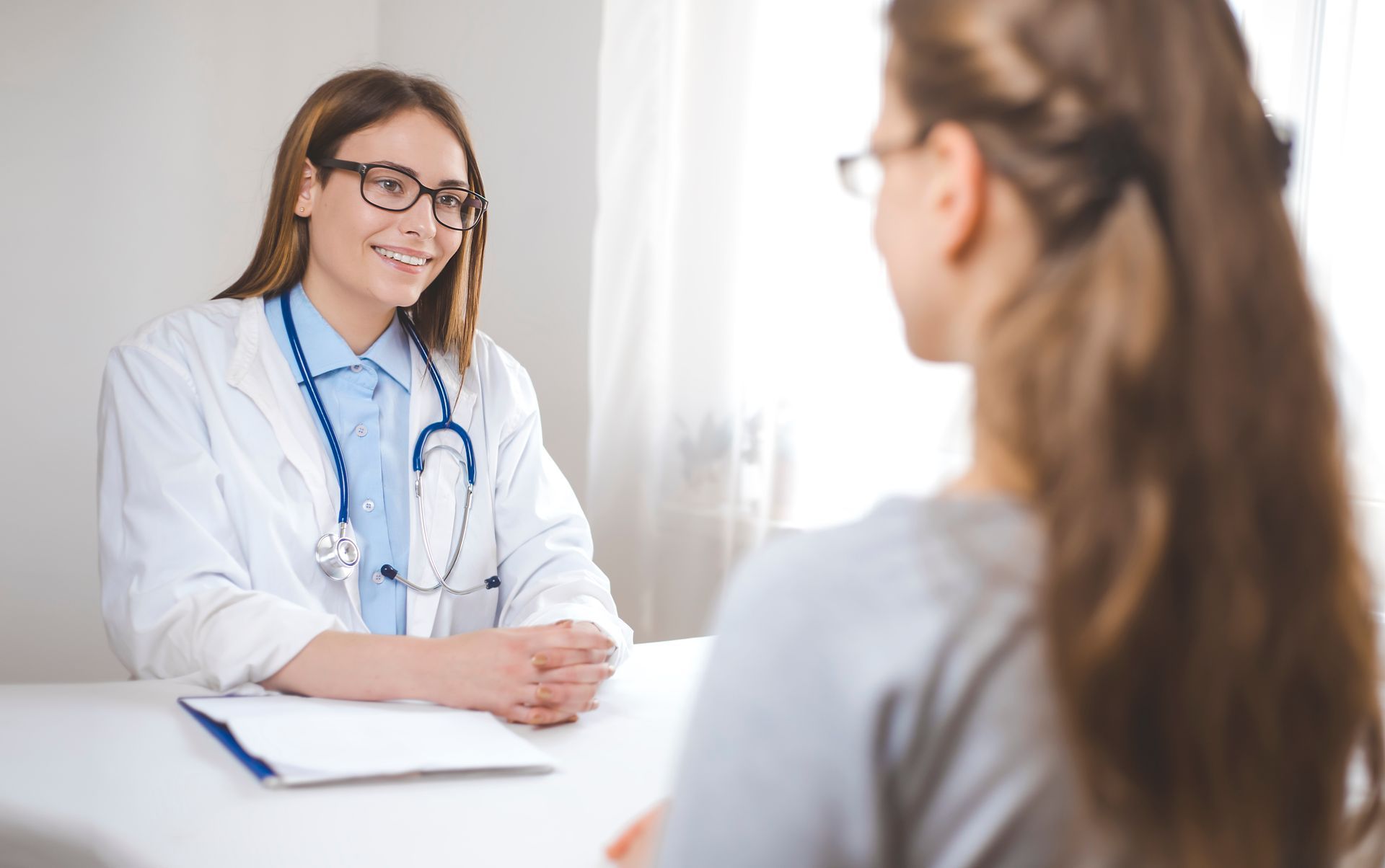 A clinician meets with a patient at a desk in a bright medical examination room. A clinician meets with a patient at a desk in a bright medical examination room.