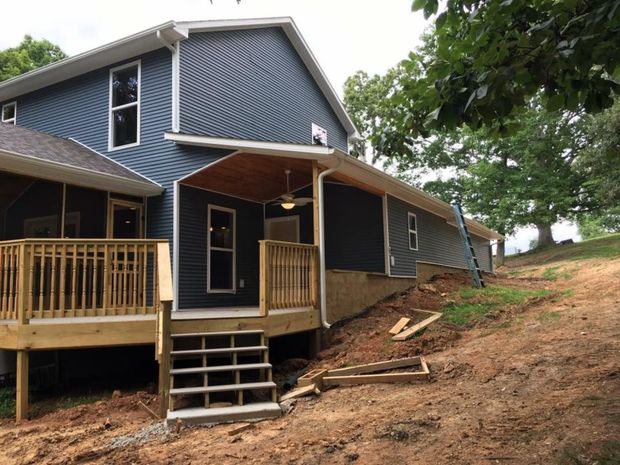 Blue house with a wooden porch on a sloped dirt lot, shown in a wooded setting