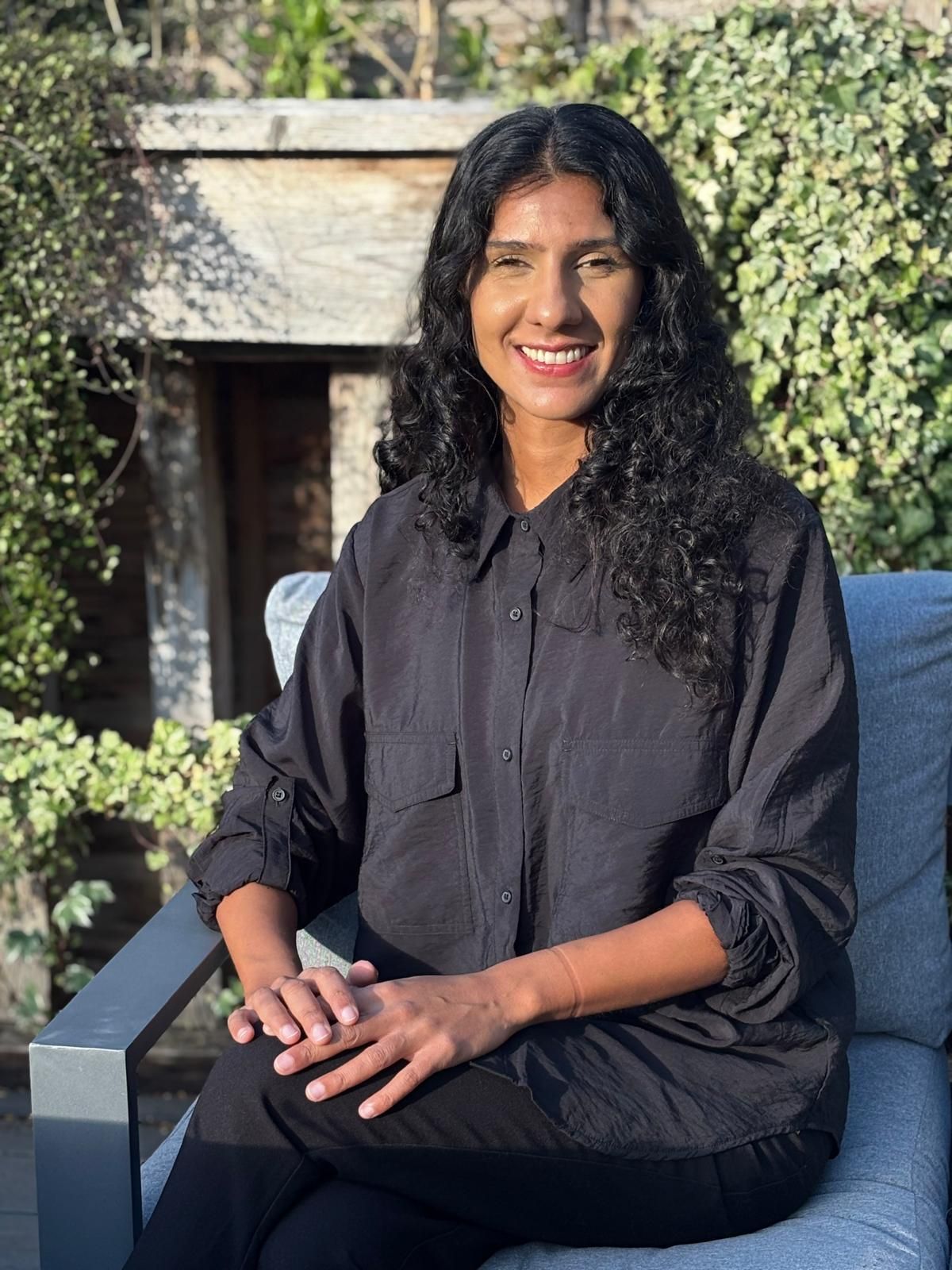 Smiling woman in black sitting outdoors on a chair with greenery and a stone wall behind her
