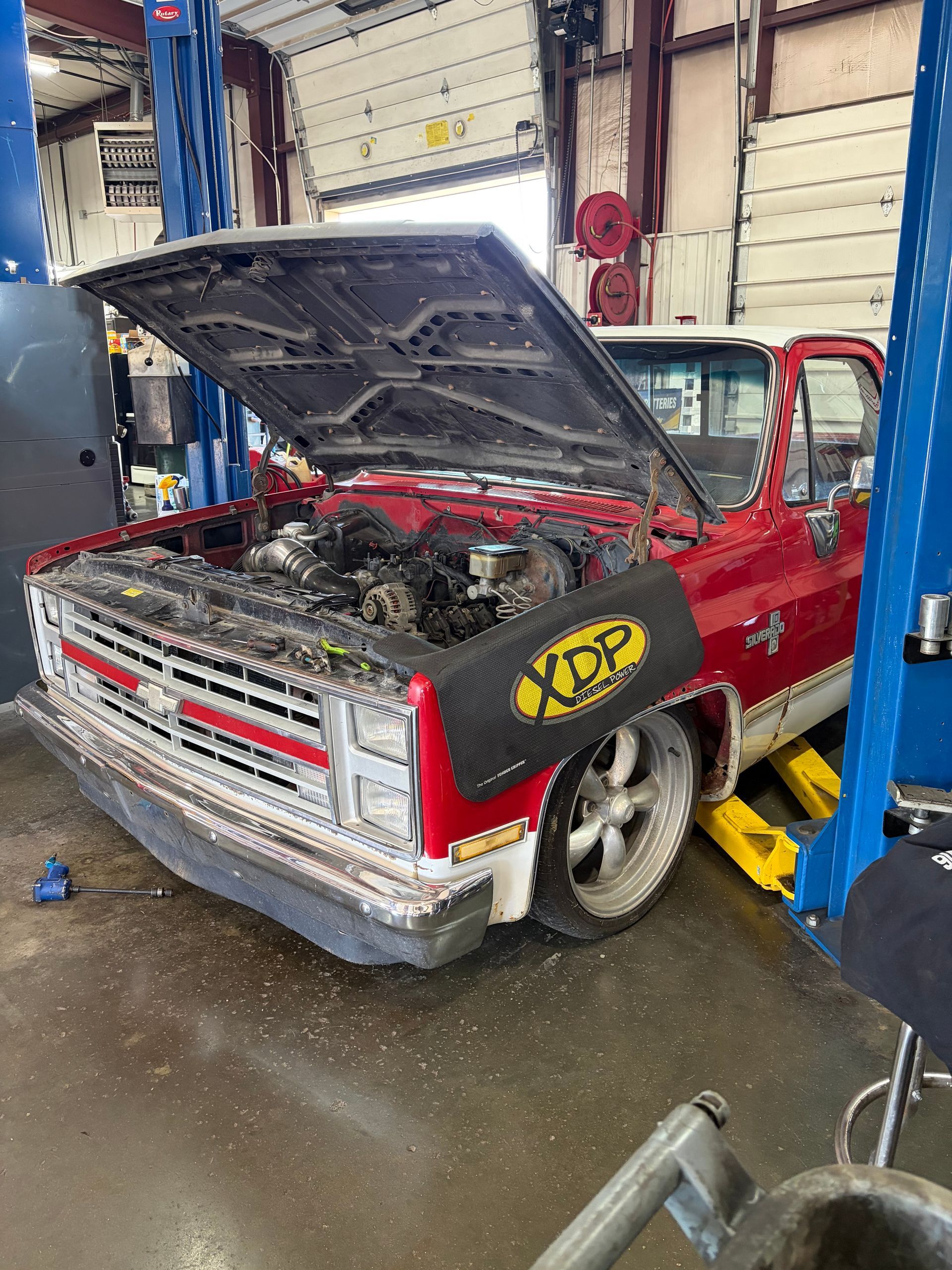 Red classic car in a garage, hood open for repair on a lift, front end partially disassembled.