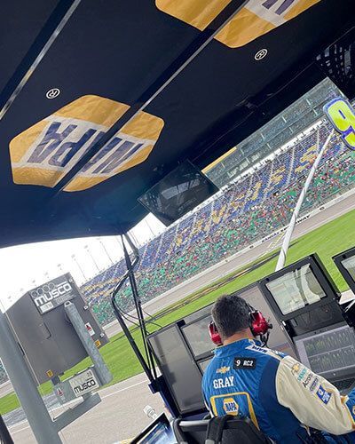 Pit crew member in blue stands beside a race car pit wall, with a packed stadium and track visible.