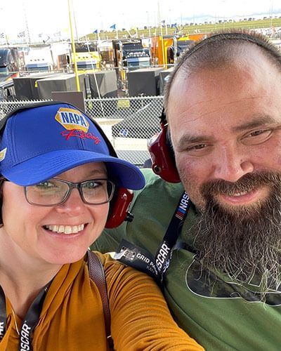 Two smiling people wearing Caps and headphones posing outdoors at a racetrack.