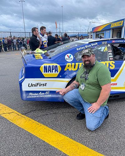 Man kneeling by blue-and-yellow race car at a track, wearing green shirt and lanyard, with spectators behind