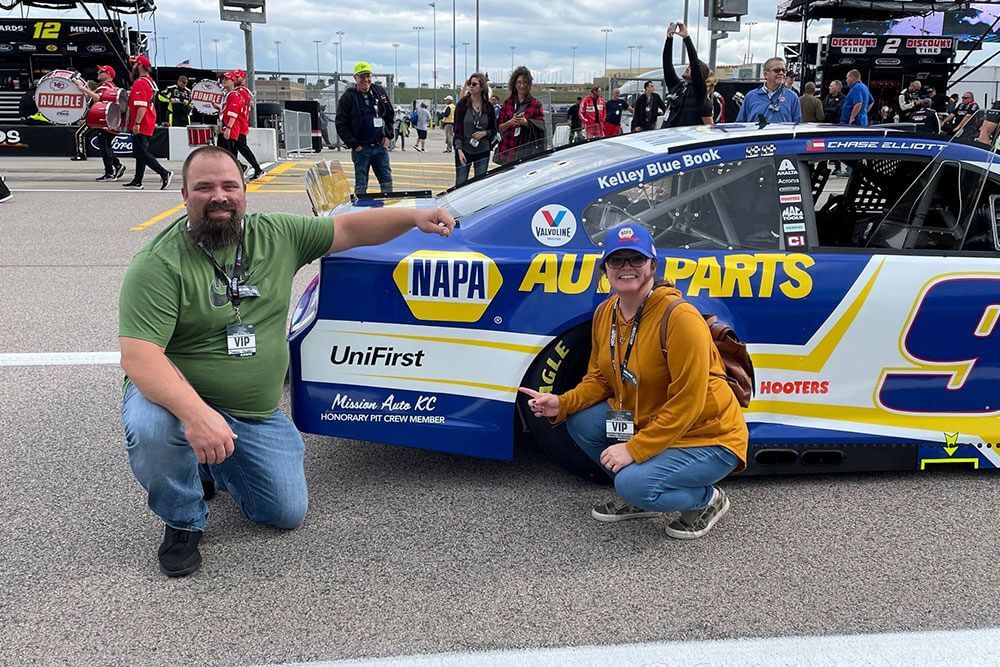 Two people kneel beside a blue NAPA Auto Parts race car at a racetrack.