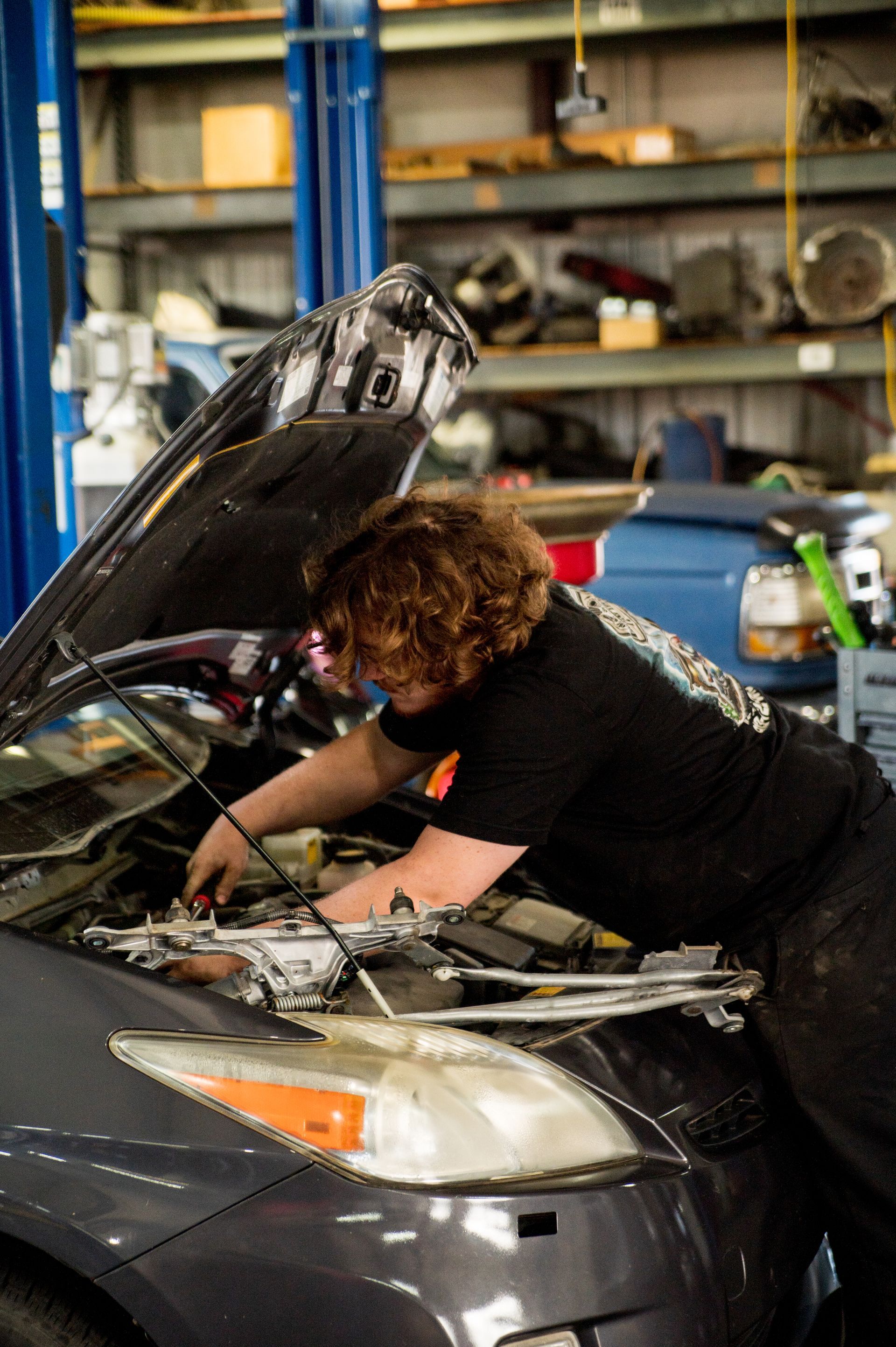 Mechanic working under a raised car hood in a garage, inspecting the engine.