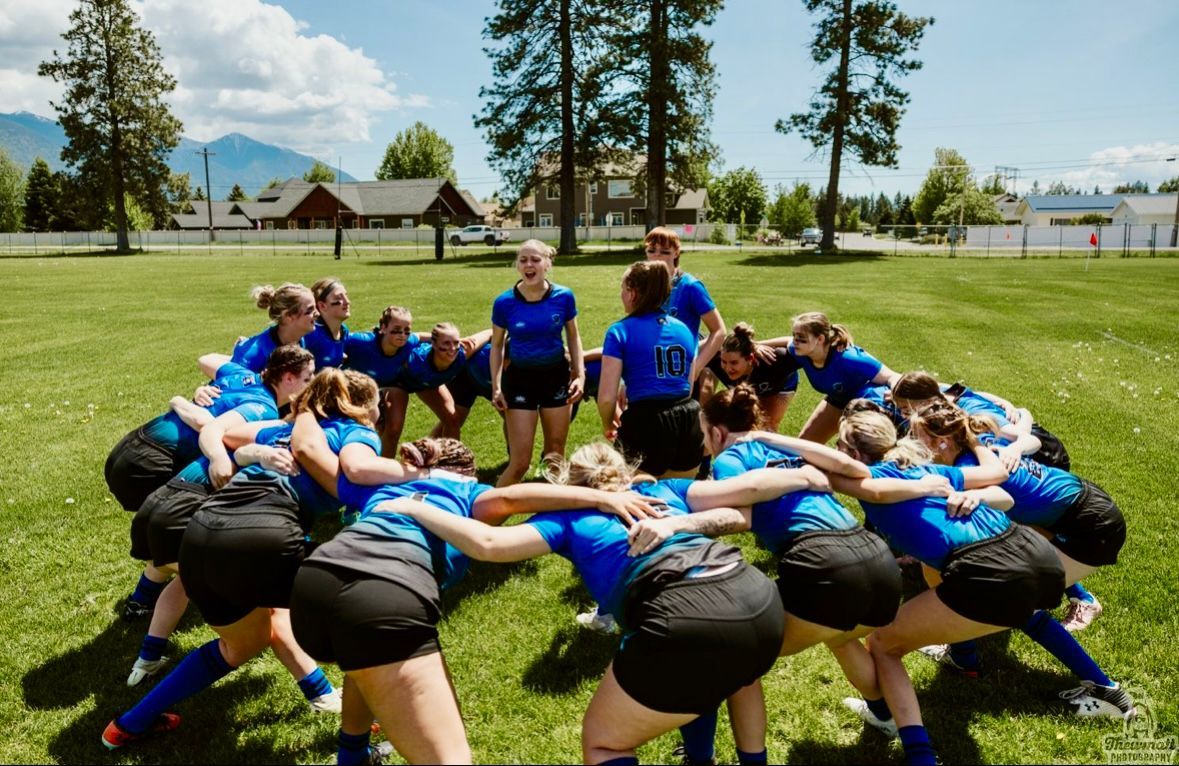 Women's rugby team in blue uniforms huddles in a circle on a grassy field, mountains in the background.
