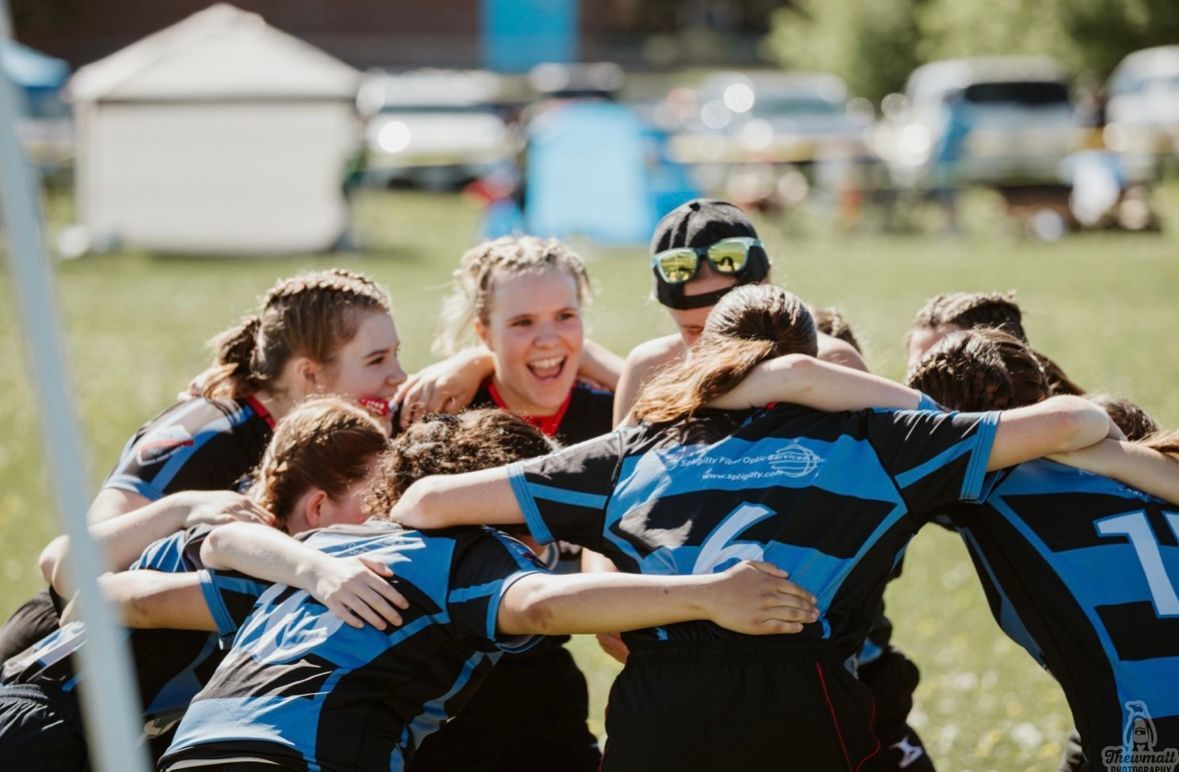 Women's rugby team huddles, laughing, arms around each other on a green field; blue and black jerseys.