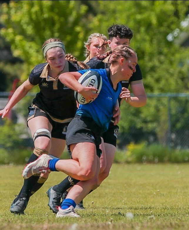 Rugby player in blue jersey runs with ball, pursued by two players in black uniforms on a grass field.