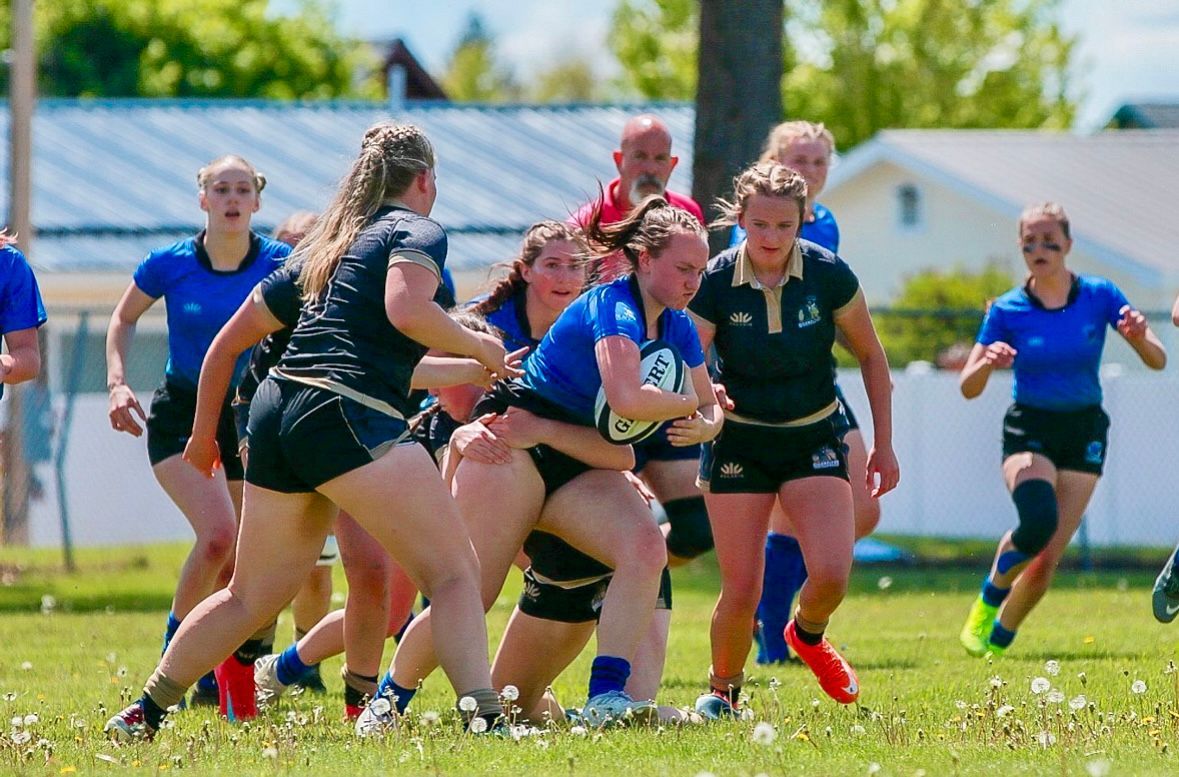 Women's rugby game. Blue and black team members tackle a player with the ball on a green field.