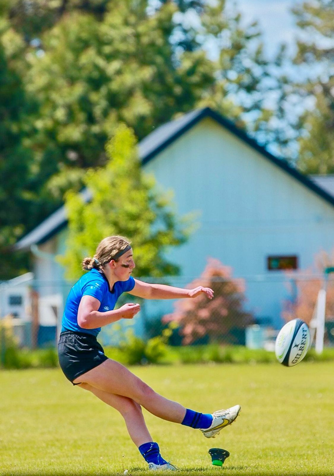 Woman in blue jersey kicks a rugby ball on a grass field. A light-colored house is in the background.