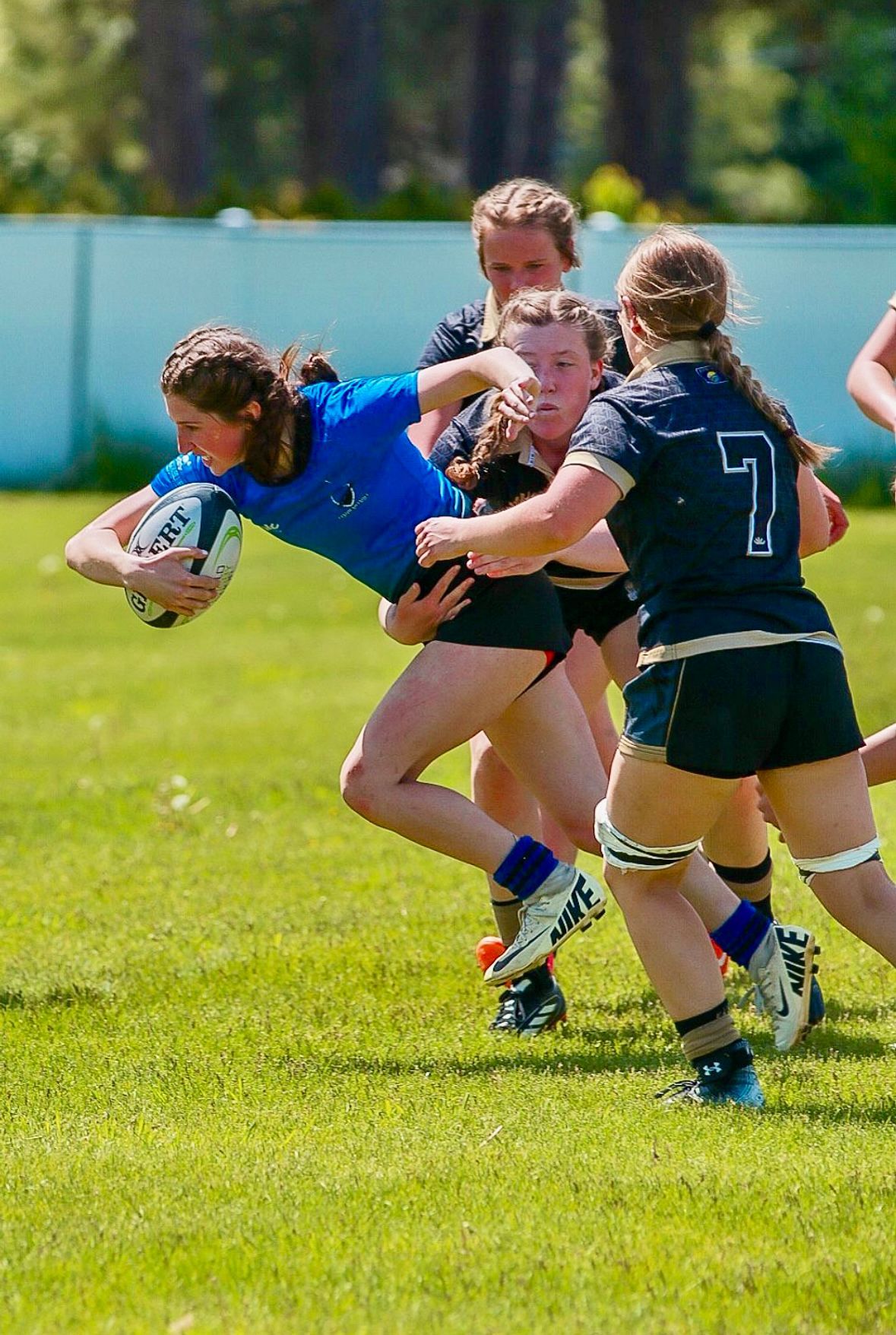 Rugby players in action: a woman in blue is tackled by two players in black on a grassy field.