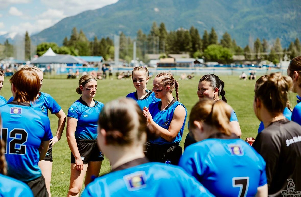 Women's rugby team huddle on a green field. Players wear blue jerseys, mountains in background.