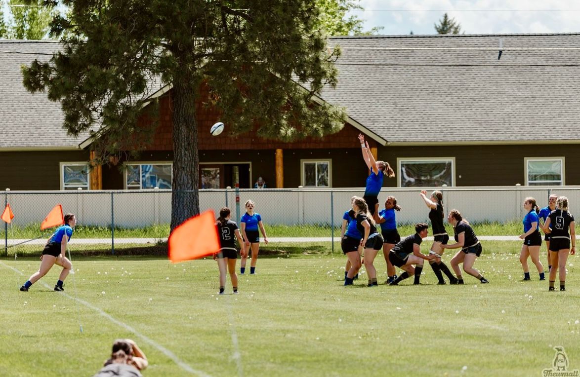 Women's rugby game; players in blue and black uniforms, jumping for ball in front of a building.