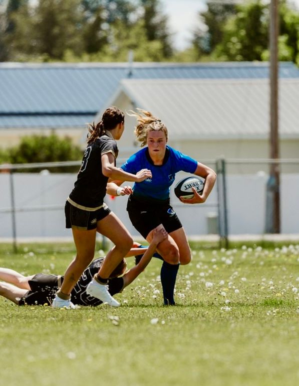 Female rugby player in blue runs with ball, pursued by black-clad player on grassy field.
