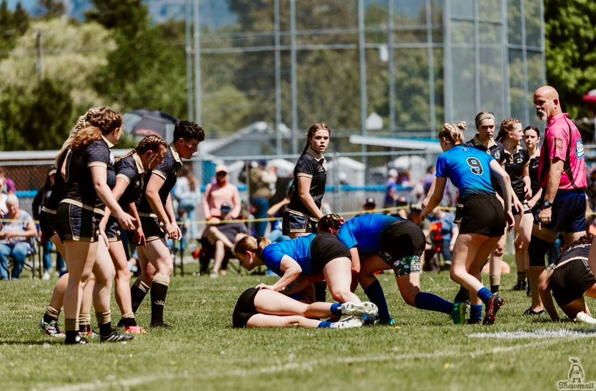 Women playing rugby on a green field. Players in black and blue jerseys collide while a referee watches.