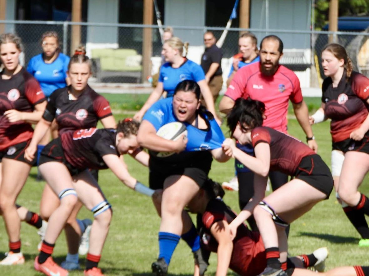 Rugby players in action, one in blue carrying the ball while being tackled by others in red and black uniforms.
