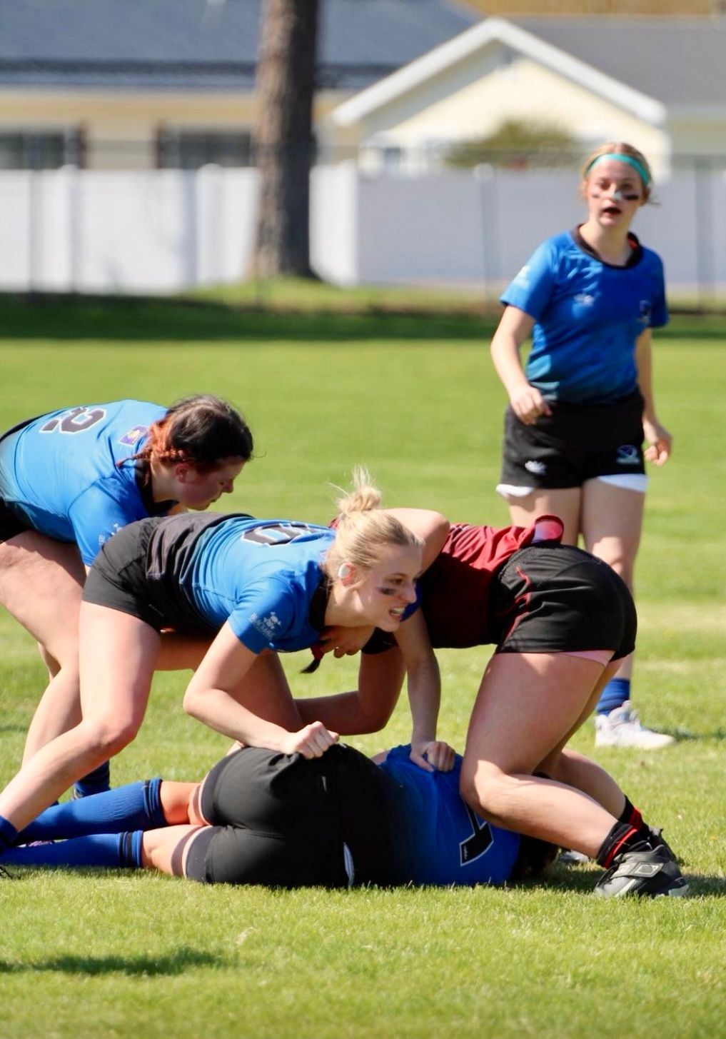 Rugby players in a scrum. One player on the ground, others crouched above. Green field, sunny day.