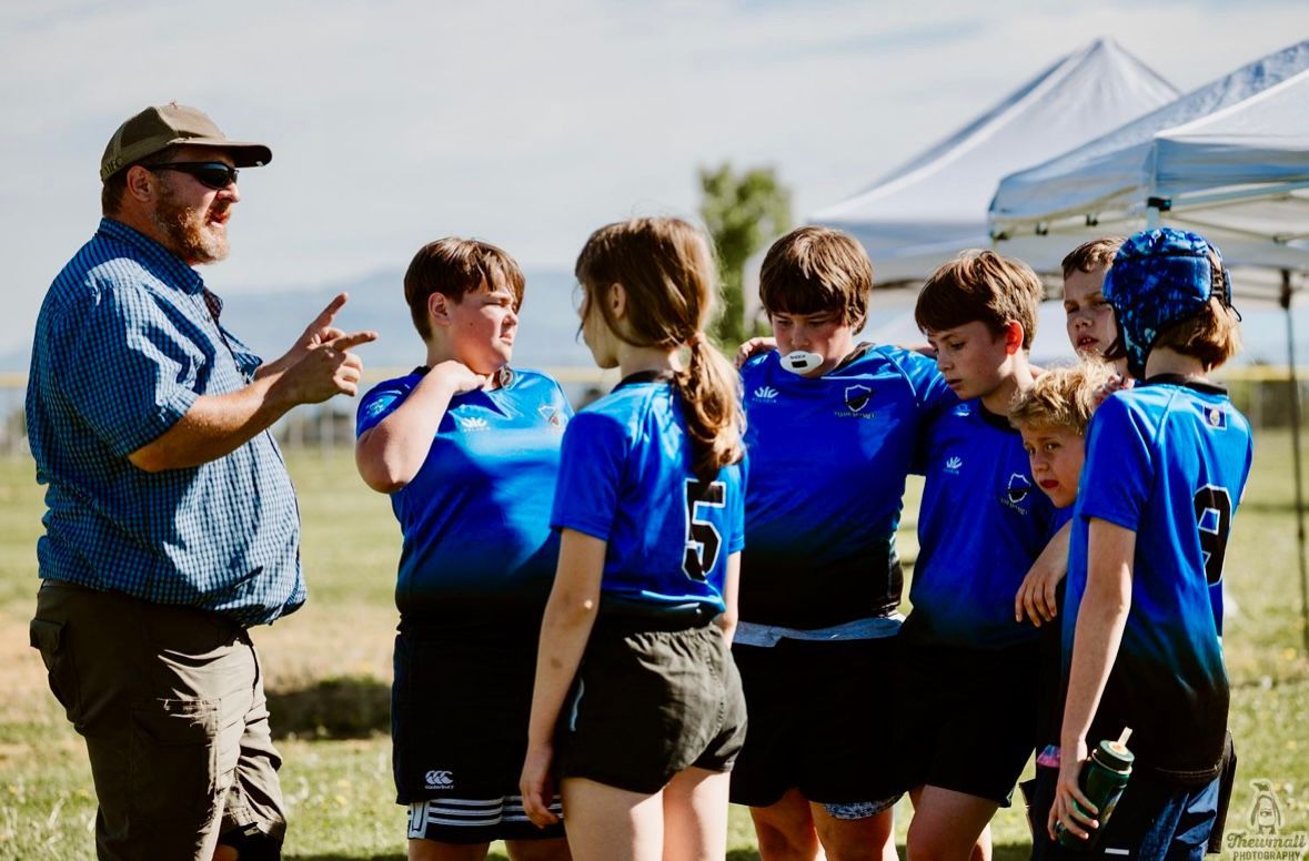 A coach giving instructions to a rugby team in blue shirts outdoors near a white tent.