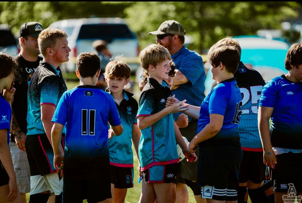 Youth rugby players in blue and black jerseys huddle on a sunny field.