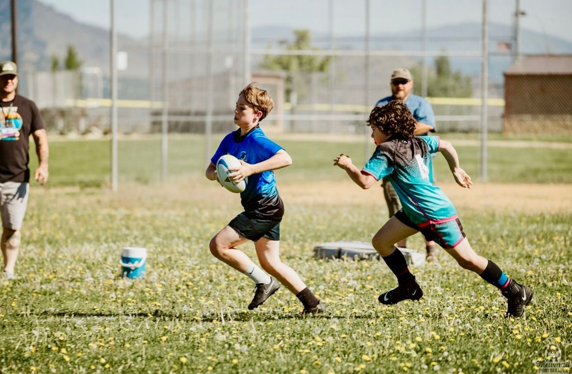 Two young boys running with a rugby ball on a green field, coaches watching in the background.