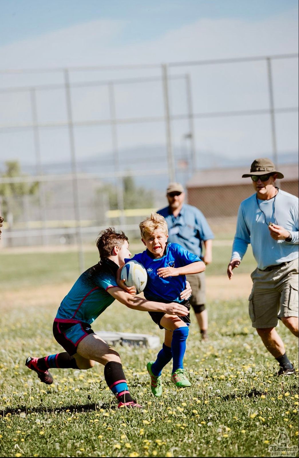Two young rugby players in action on a sunny field, one passing the ball, coaches watching.