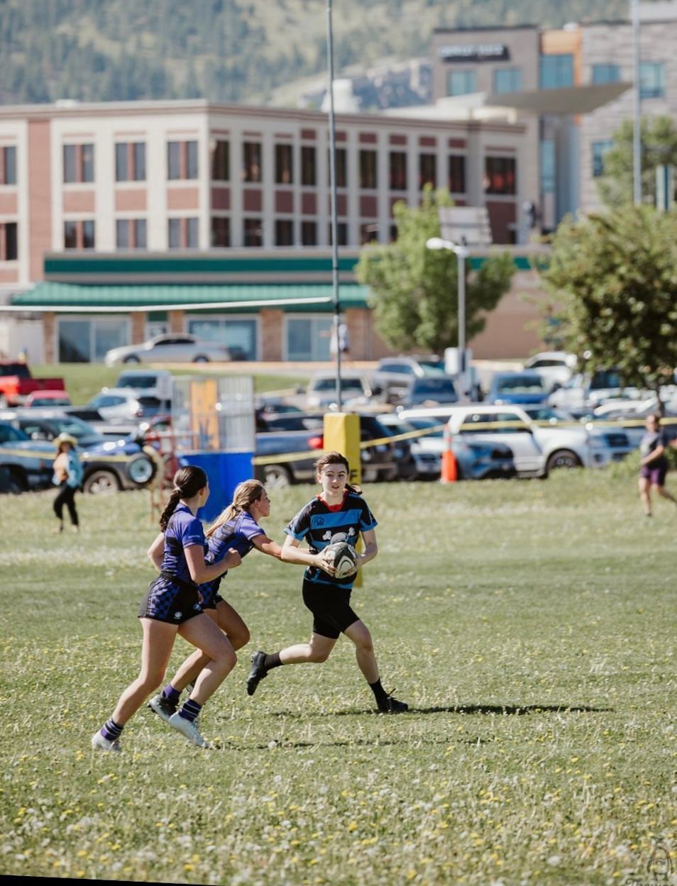 Rugby player runs with ball, pursued by two women in blue. Field in front of buildings and parked cars.