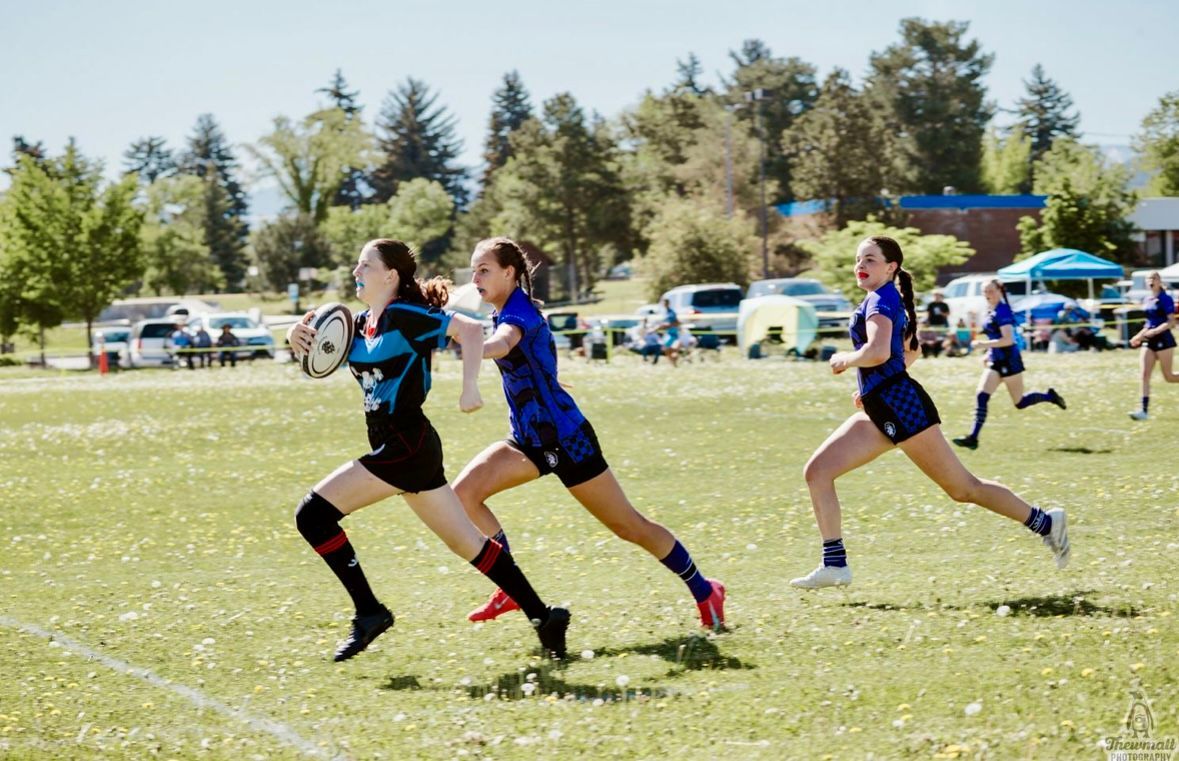 Women's rugby match: three players running on a grassy field. One holds the ball; others follow. Sunny, outdoor setting.