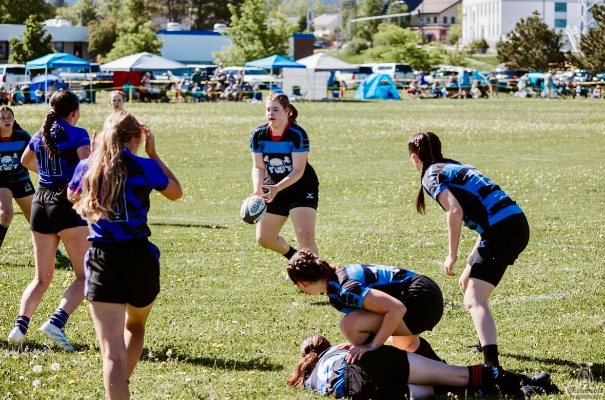 Women's rugby game in a field. Players in blue and black uniforms, one about to pass the ball.