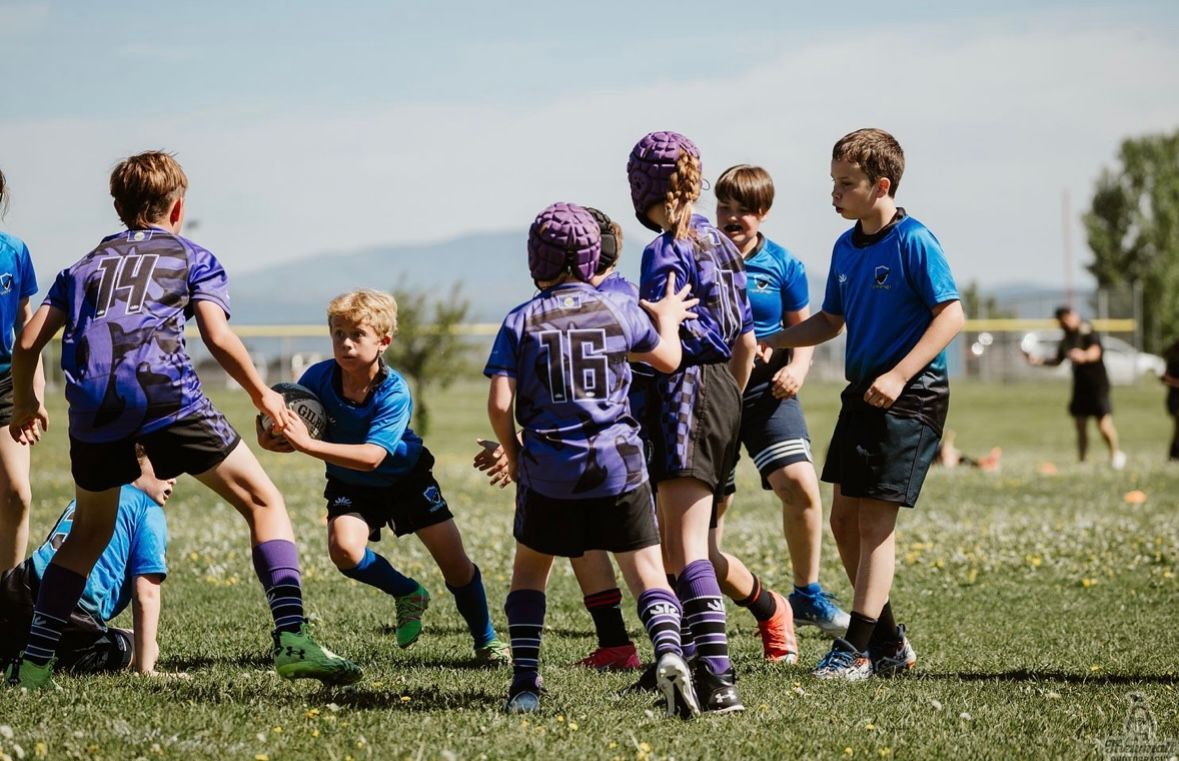 Children in purple and blue jerseys play rugby on a grassy field.