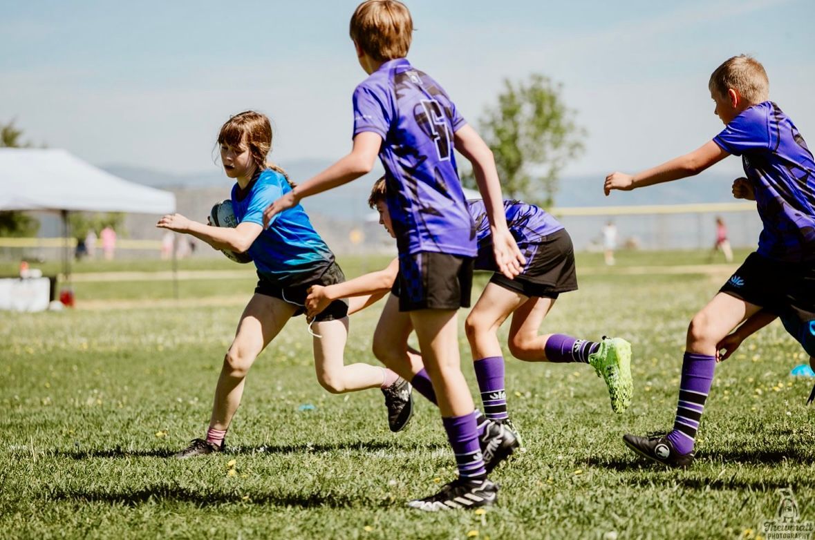 Kids in blue and purple jerseys run during a rugby game on a grassy field.