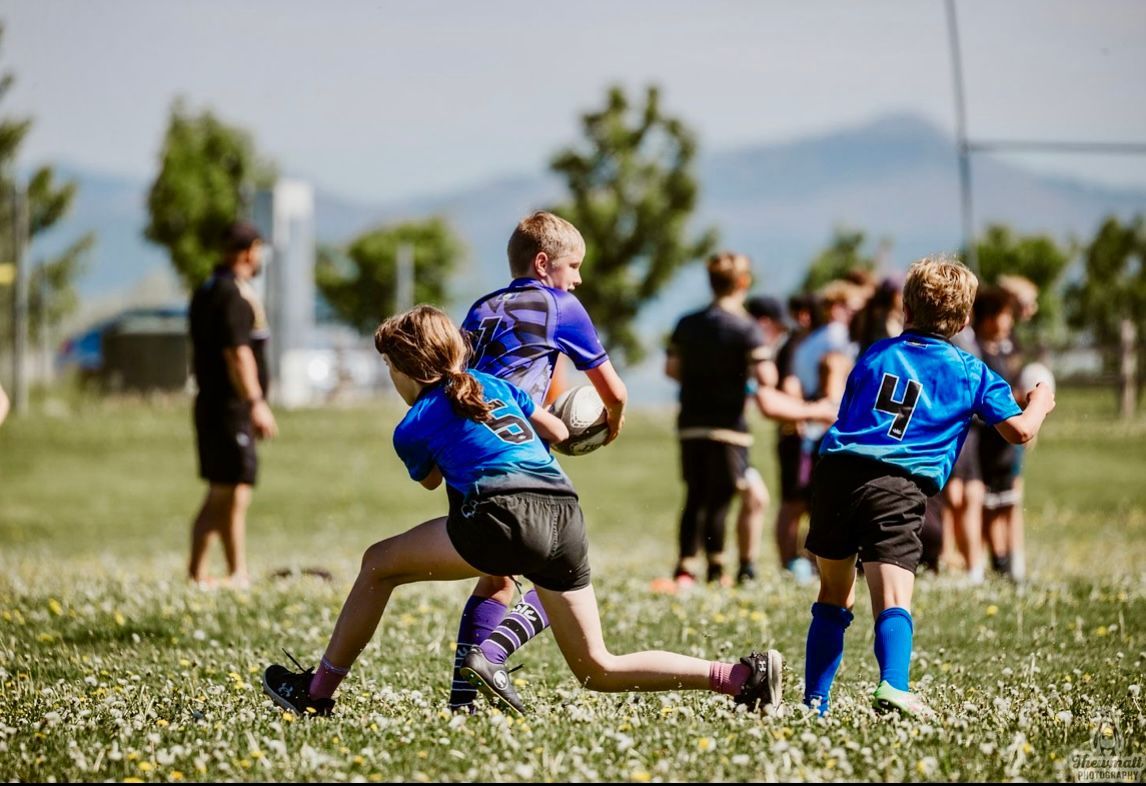 Kids playing rugby in a grassy field. One player runs with the ball, others chase. Mountains in the background.