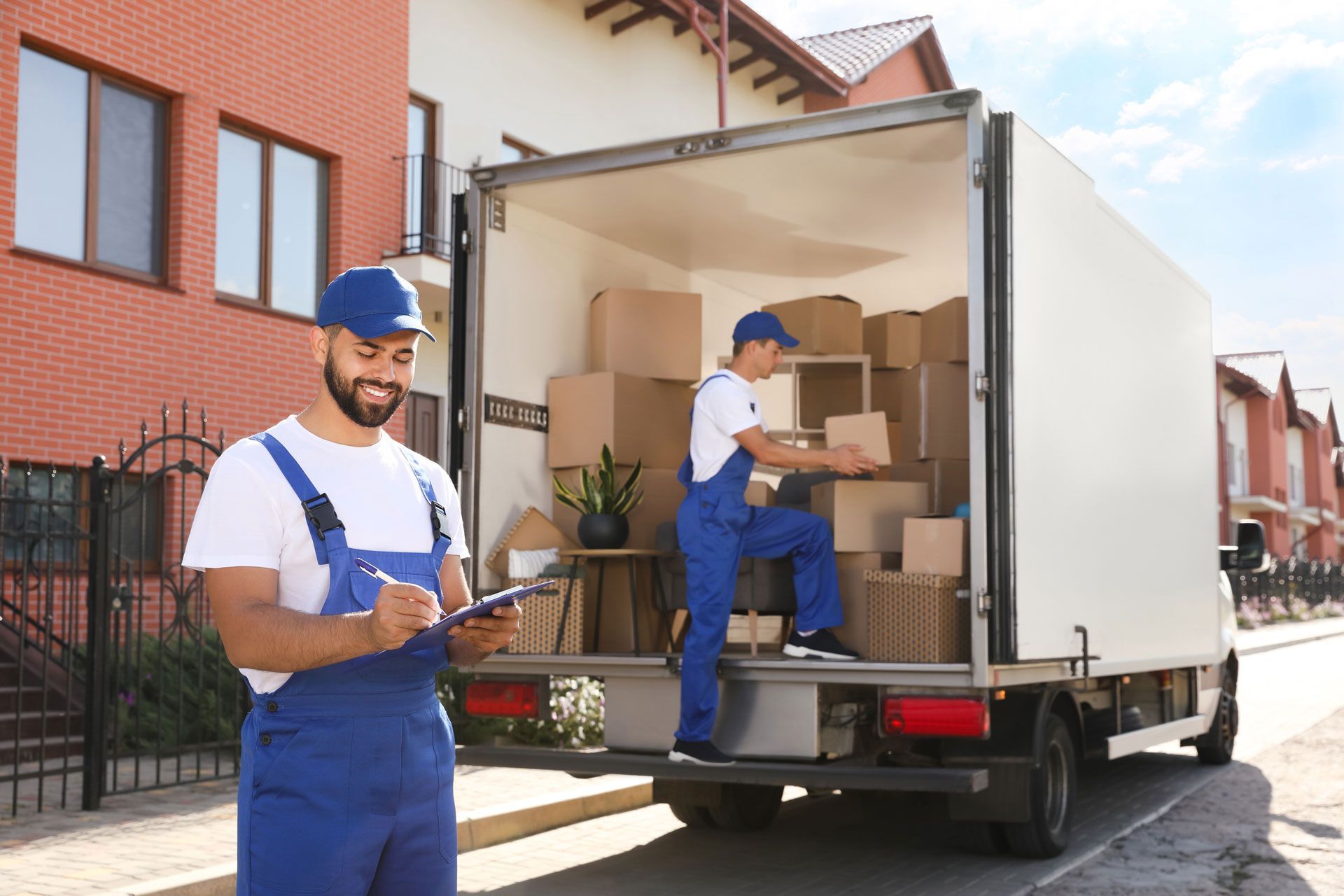 Two movers loading boxes into a truck parked in front of a house. One writes on a clipboard, the other stacks boxes.