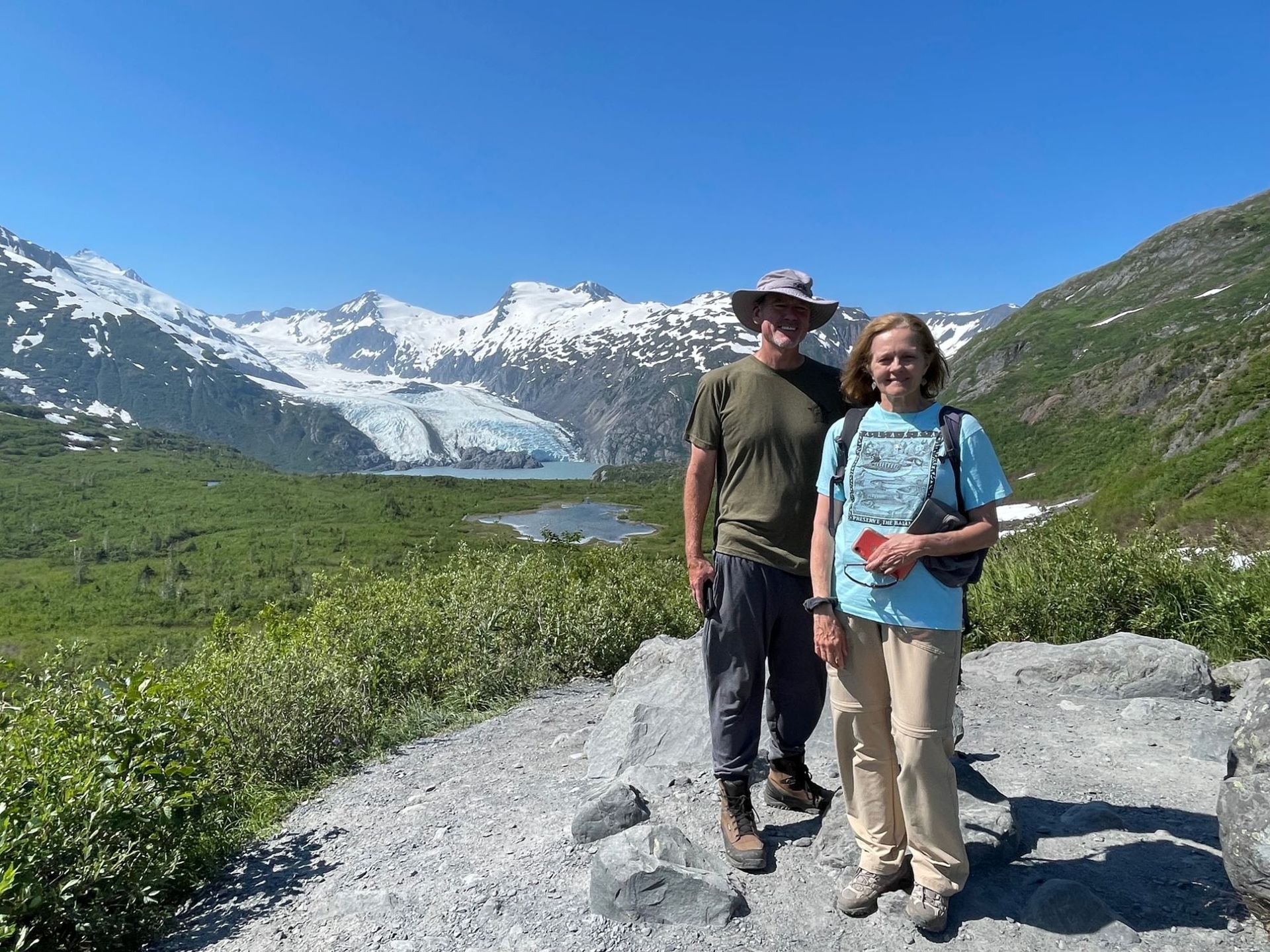 Two hikers standing on a mountain trail with snow-capped peaks and a lake behind them
