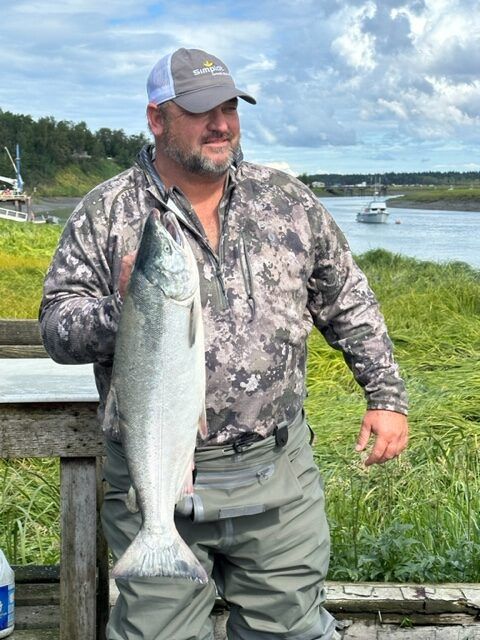 Man holding a large silver fish beside a wooden railing with a lake and sailboats in the background
