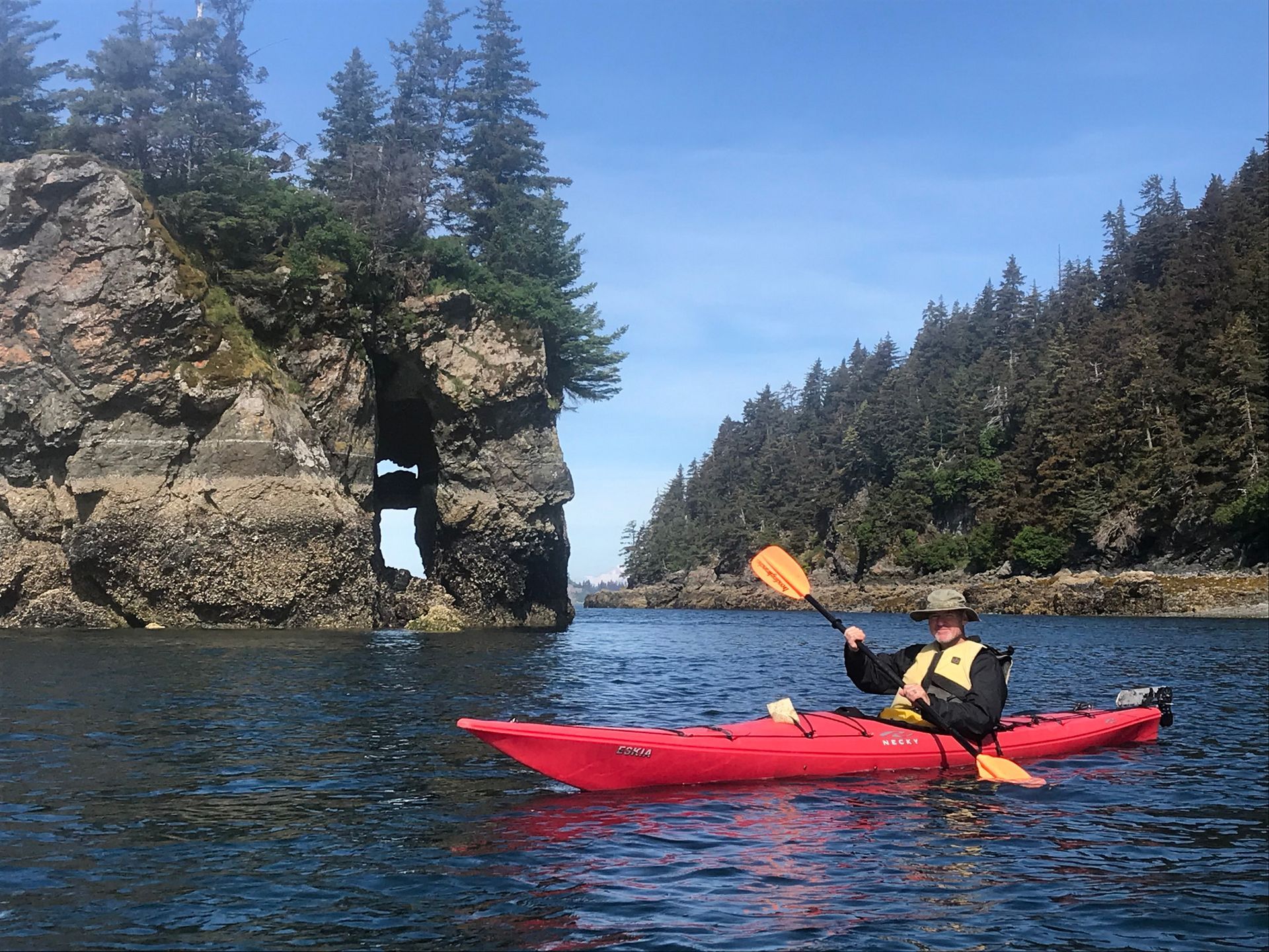 Red kayak with paddler on calm water beside rocky, tree-covered cliffs and a sea arch.